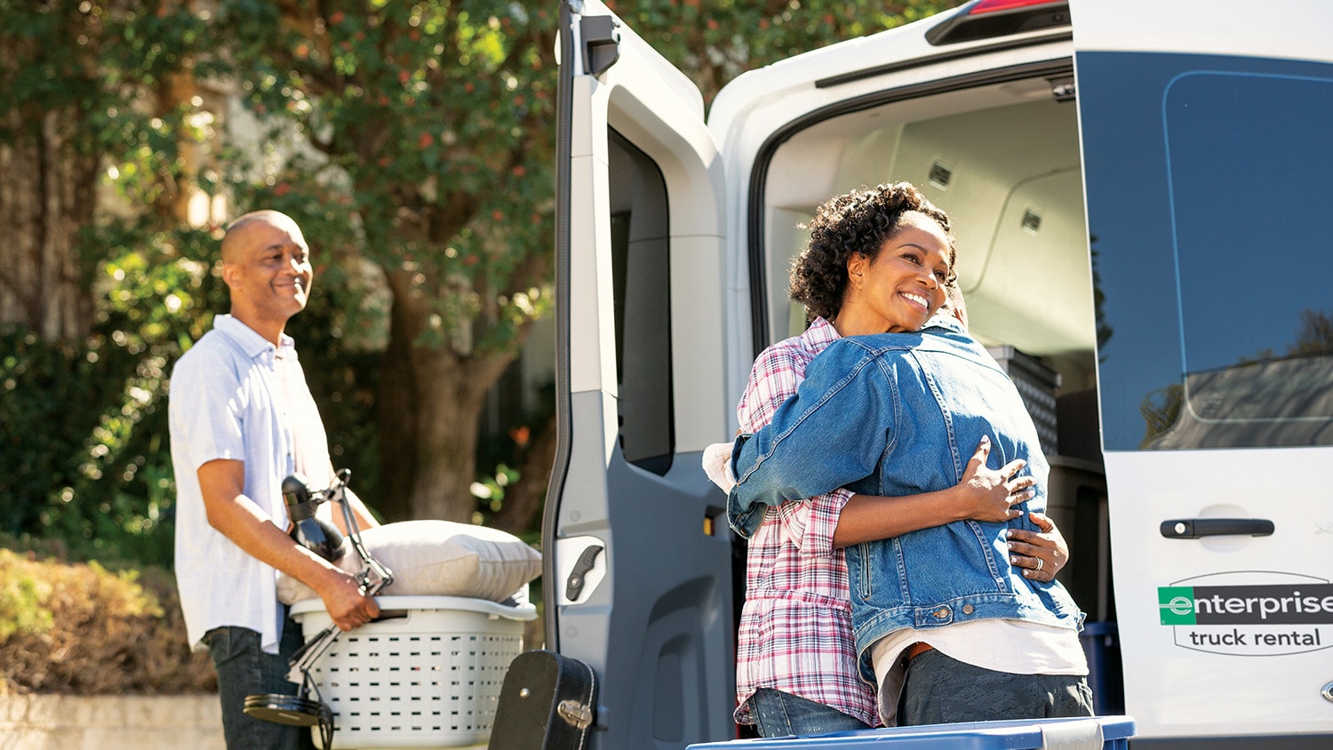 Woman is hugging a younger man outside the back of a moving van, whie older man is watching 